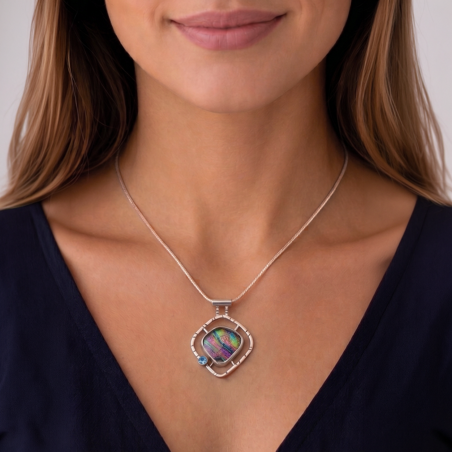 A close-up of a woman wearing a silver-toned necklace with a square pendant featuring an iridescent, multicolored inlay and a small blue accent stone.