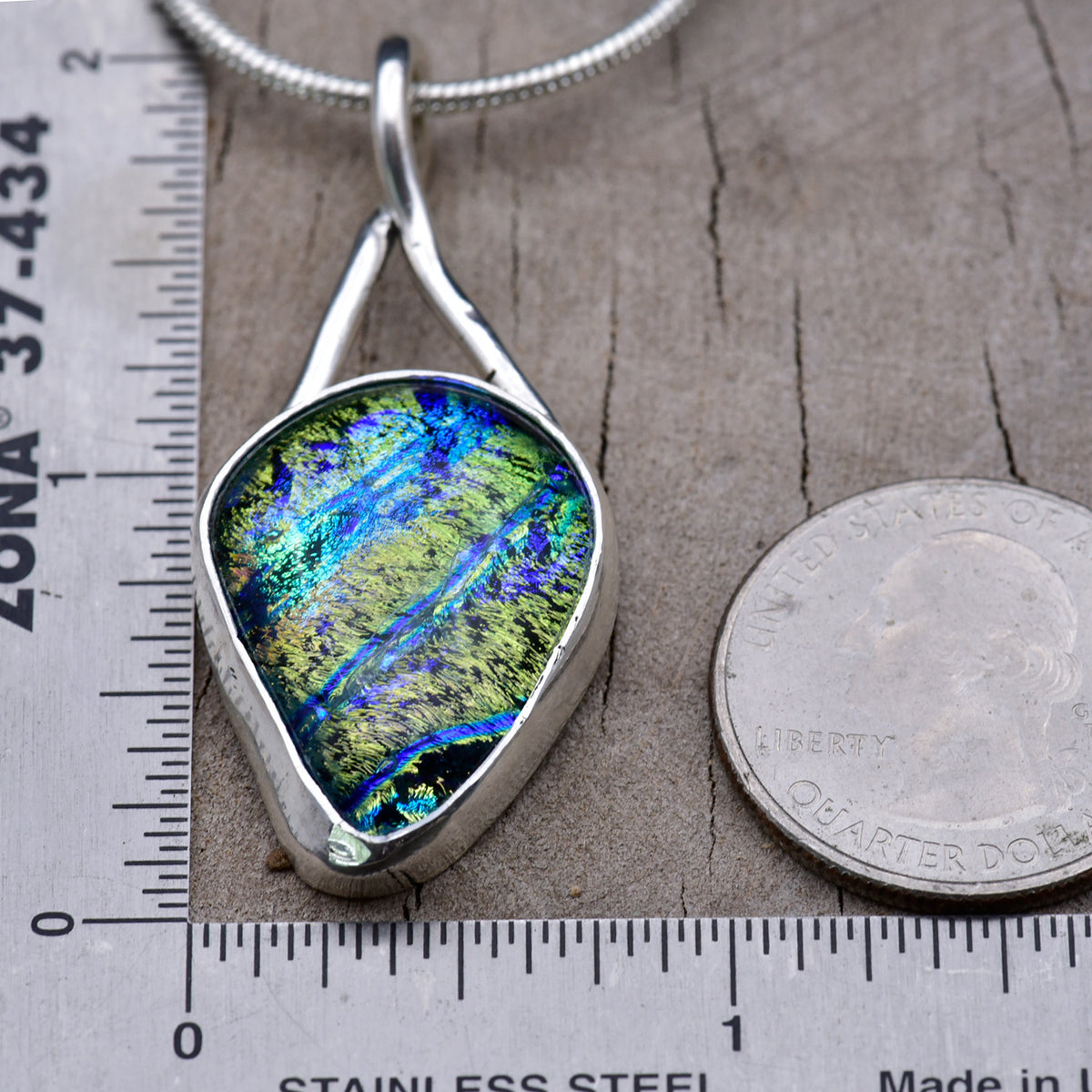 Green and cobalt dichroic glass teardrop pendant beside a ruler and a quarter for scale, measuring about 2 inches tall and 1 inch at its widest point.