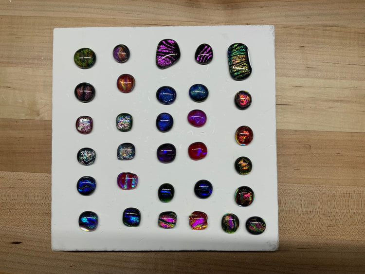 Around thirty fused dichroic glass cabochons in many colors, arranged on a white kiln shelf.