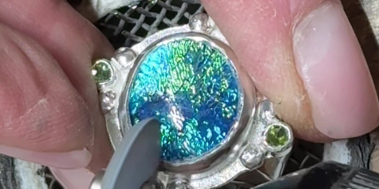 Hands setting a teal and green dichroic glass ring with peridot accents, using a burnisher at the jeweler's bench.