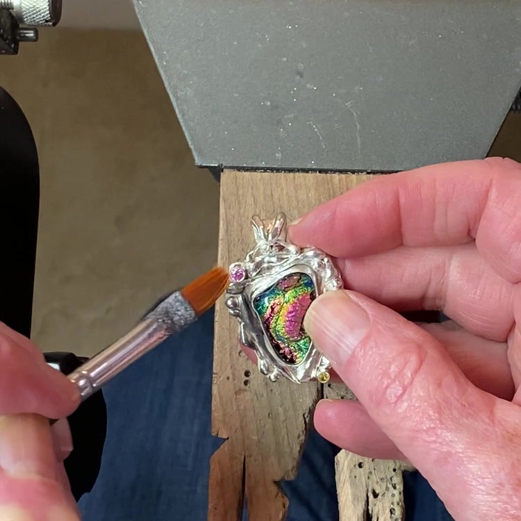 Hands at a jeweler's bench applying patina with a fine brush to a handmade silver pendant set with rainbow dichroic fused glass.