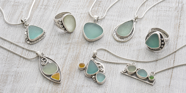 Sea glass pendants and rings in sterling silver and mixed metals, arranged on weathered pale wood.