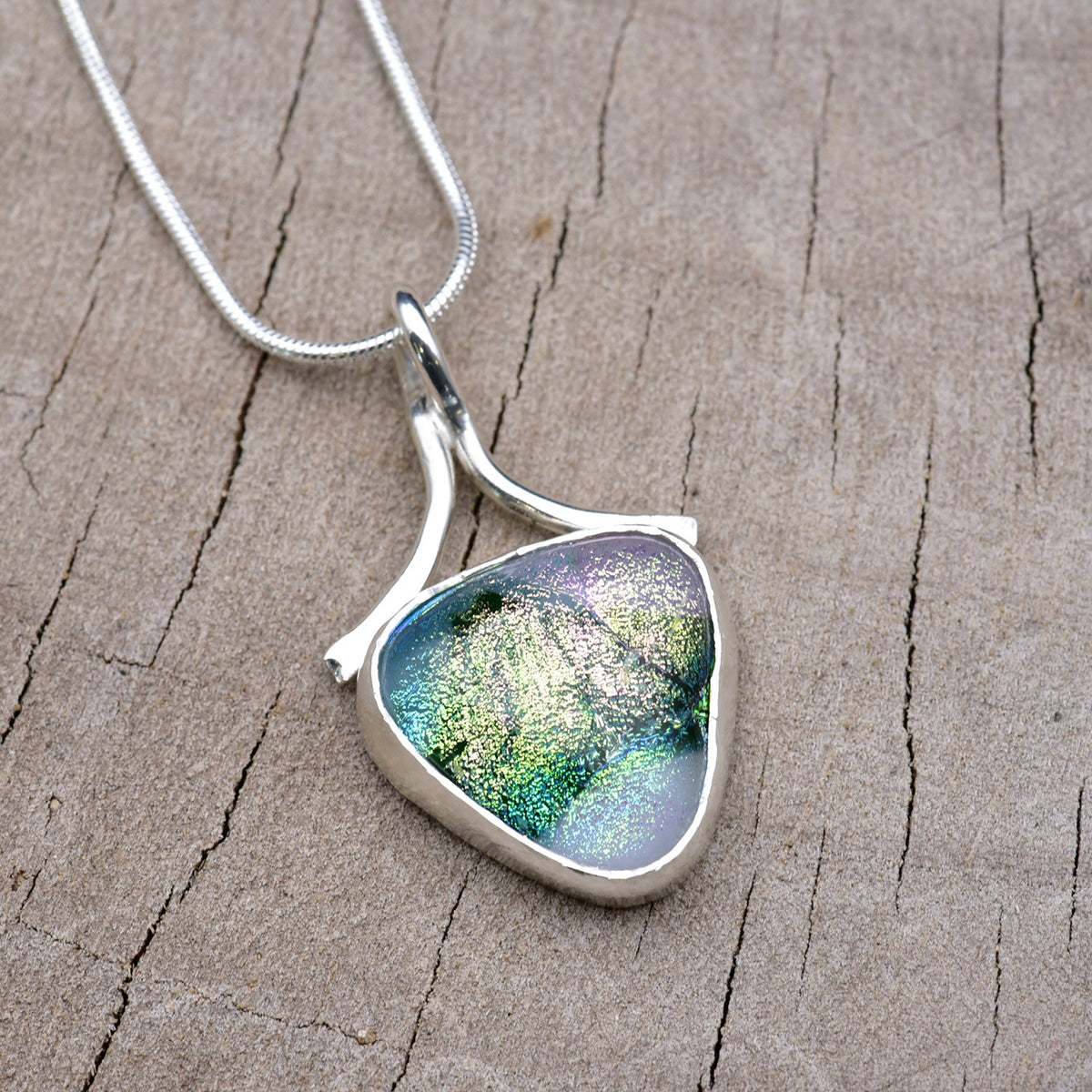 Handmade shield-shaped pendant necklace with a dichroic glass cabochon shading from pearly lilac and silver into emerald and teal, in an Argentium sterling silver setting with a ribbon bail, on a silver snake chain.