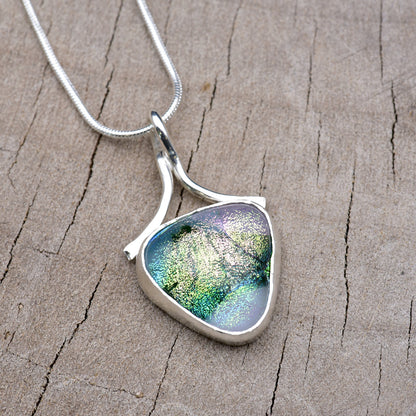 Handmade shield-shaped pendant necklace with a dichroic glass cabochon shading from pearly lilac and silver into emerald and teal, in an Argentium sterling silver setting with a ribbon bail, on a silver snake chain.
