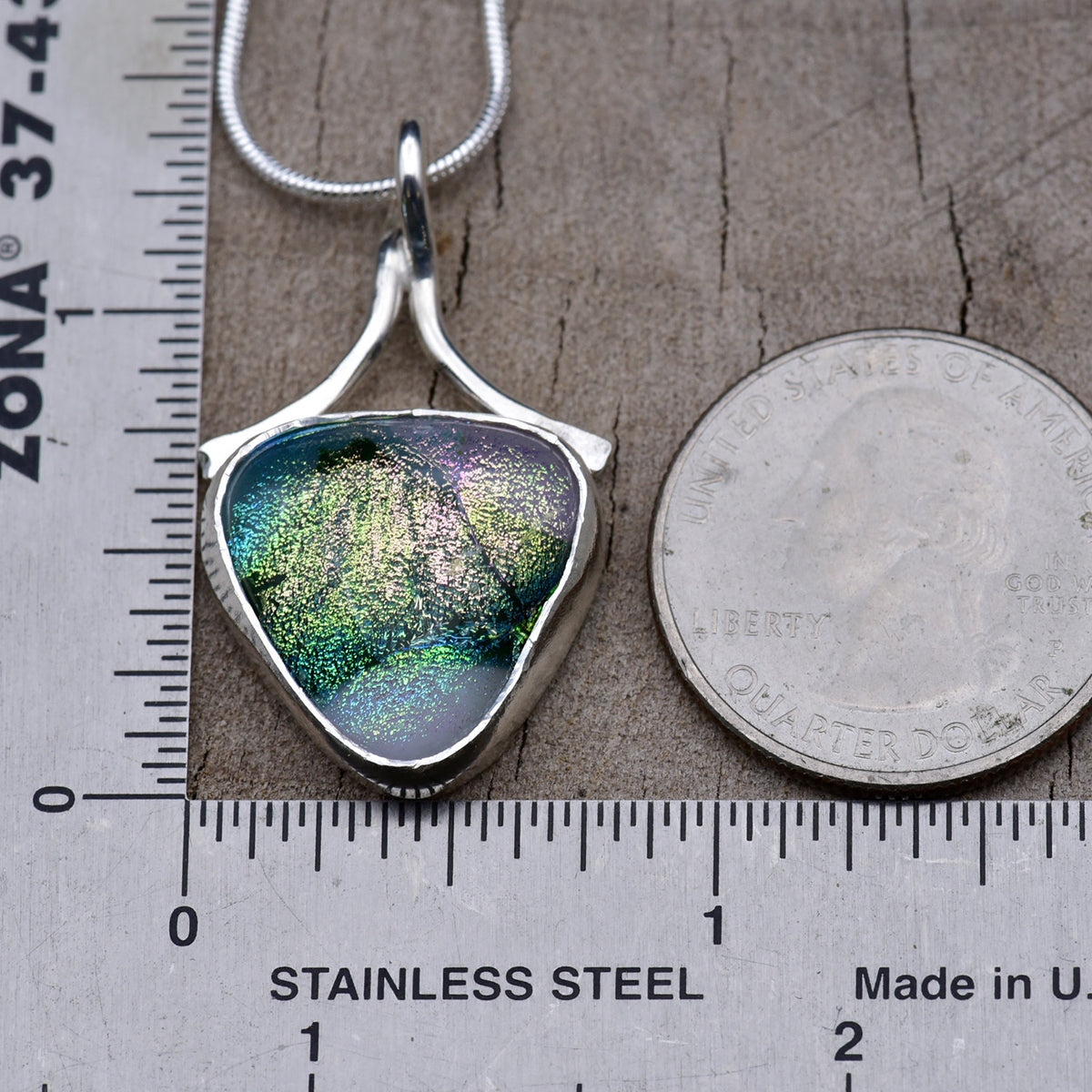 Lilac and emerald dichroic glass shield pendant beside a ruler and a quarter for scale, about 1¼ inches long and ¾ inch wide.
