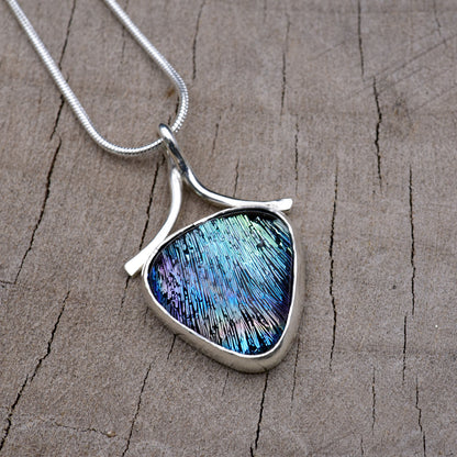 Handmade shield-shaped pendant necklace with a peacock blue and teal dichroic glass cabochon streaked with violet and magenta, in an Argentium sterling silver setting with a ribbon bail, on a silver snake chain.