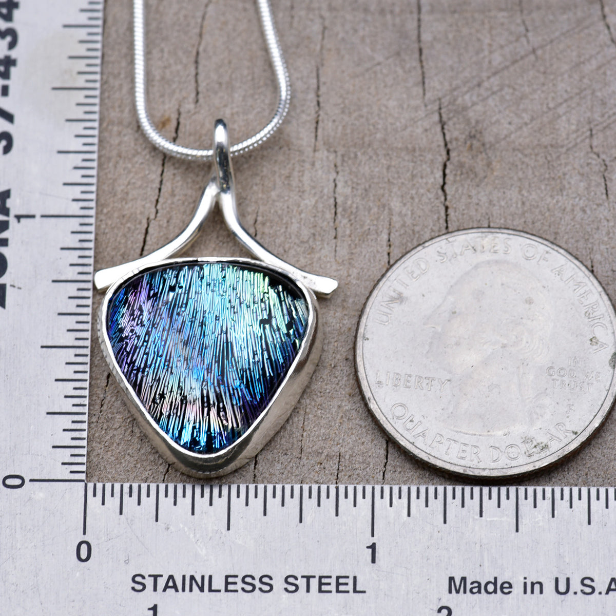 Peacock blue and violet dichroic glass shield pendant beside a ruler and a quarter for scale, about 1¼ inches long and ¾ inch wide.