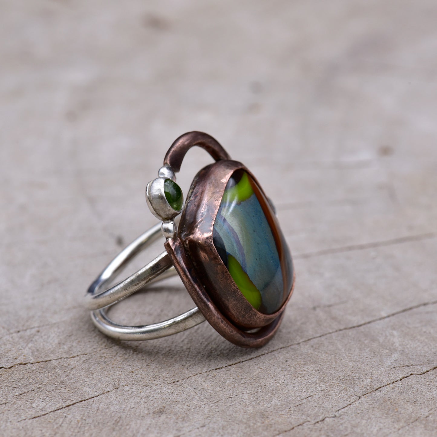 Copper and silver ring with colorful stones on a textured surface.