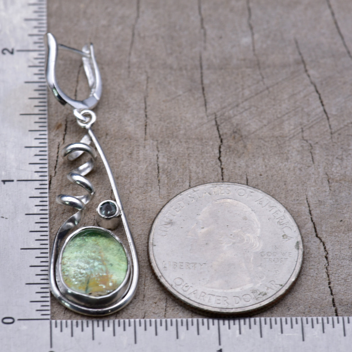 Sea green dichroic glass earring beside a ruler and a quarter for scale, measuring about 2 inches long