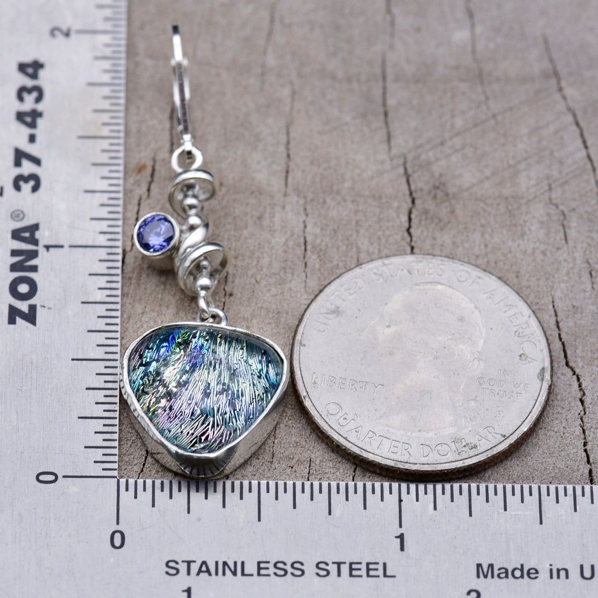 Teal and lavender dichroic glass earring beside a ruler and a quarter for scale, measuring about 1¾ inches long