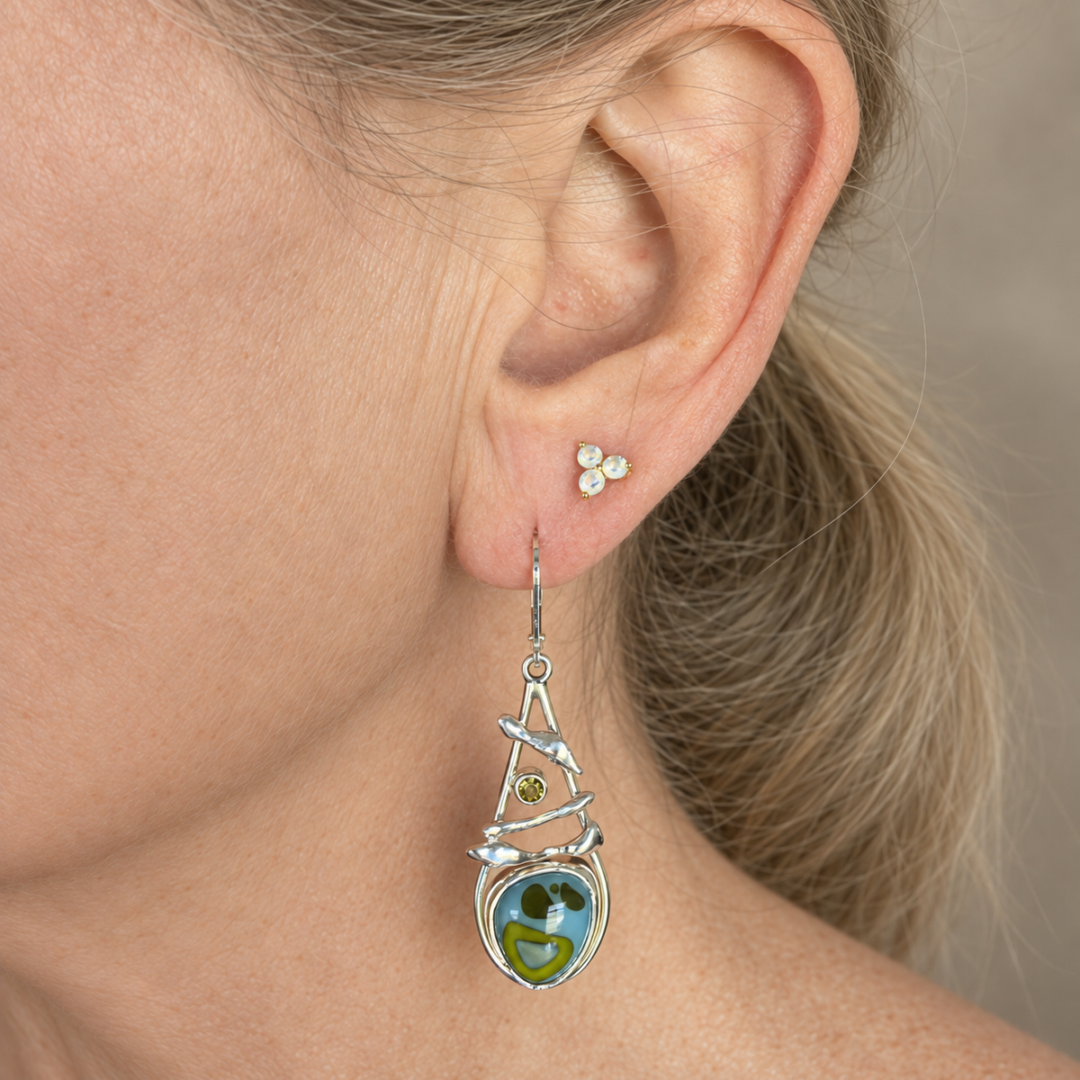 Sky blue and lime green fused glass earring worn on the ear, showing the sculpted silver branches and green stone above the glass