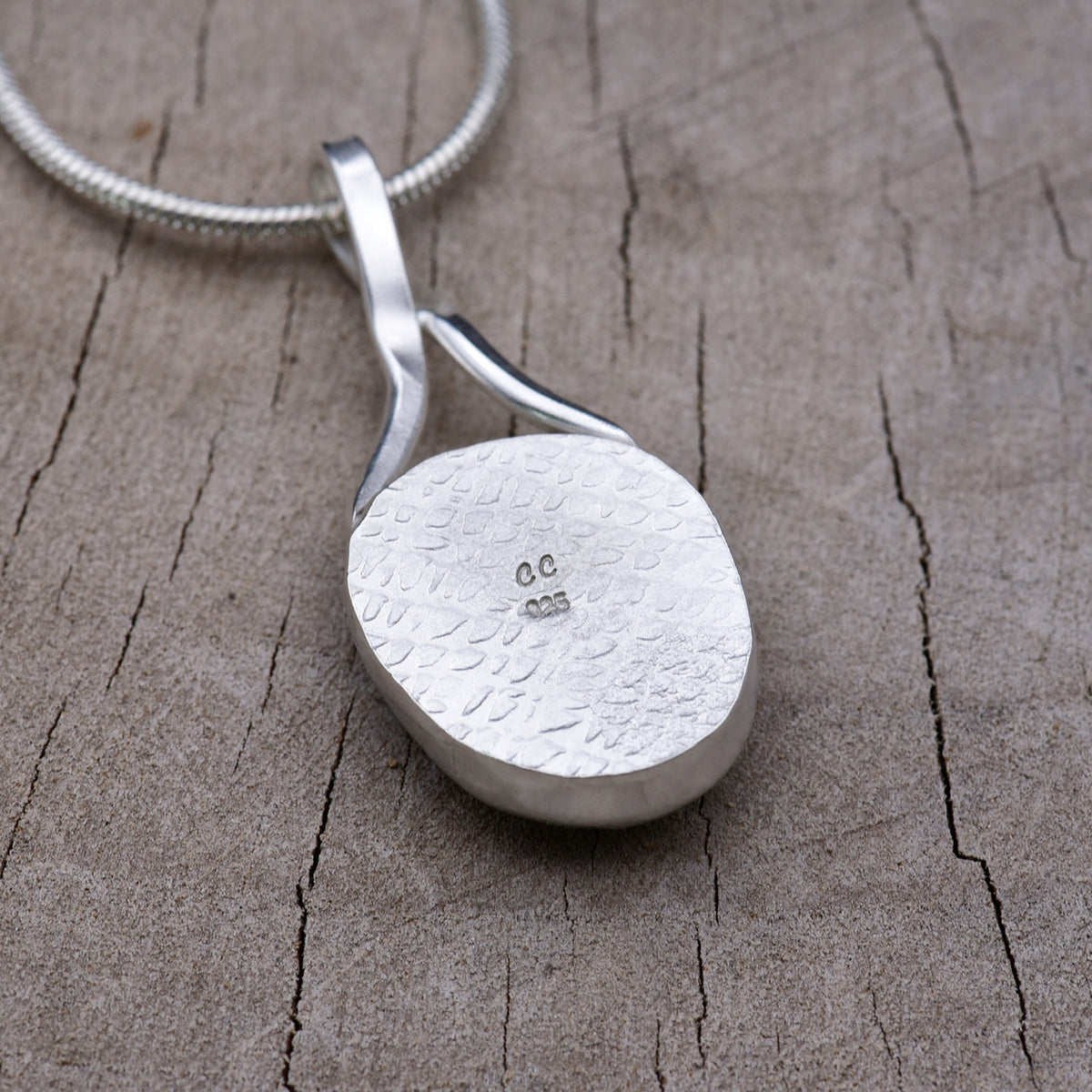 Hand-stamped and oxidized silver back of the turquoise dichroic pendant, showing the pebbled texture and the CC maker's mark above the 925 hallmark.