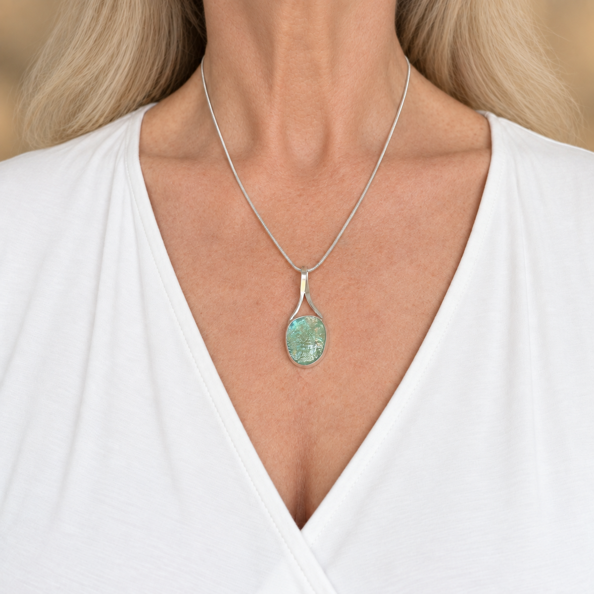 Turquoise dichroic glass pendant worn at the collarbone on a silver-plated snake chain over a white V-neck top.