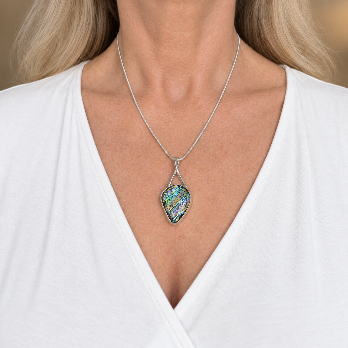 Green and blue dichroic glass teardrop pendant worn at the collarbone on a silver-plated snake chain over a white V-neck top.