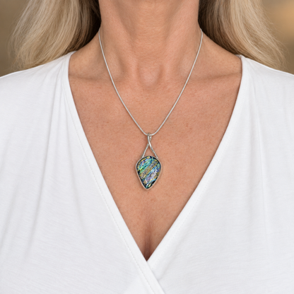 Green and blue dichroic glass teardrop pendant worn at the collarbone on a silver-plated snake chain over a white V-neck top.