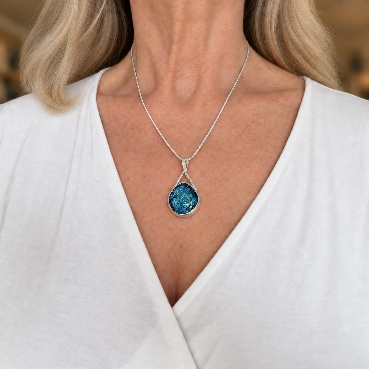Blue dichroic glass pendant worn on a snake chain, resting just below the collarbone with a white V-neck top.