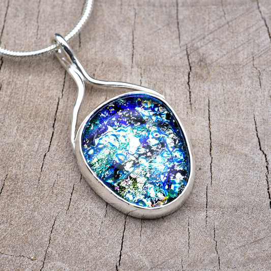 Handmade pendant necklace with a sapphire blue dichroic glass cabochon flecked with silver-white and sea green, in an Argentium sterling silver setting with a ribbon bail, on a silver snake chain.